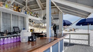Un bar en la playa con una barra de madera, estantes con botellas, una máquina de café y sombrillas en la terraza.