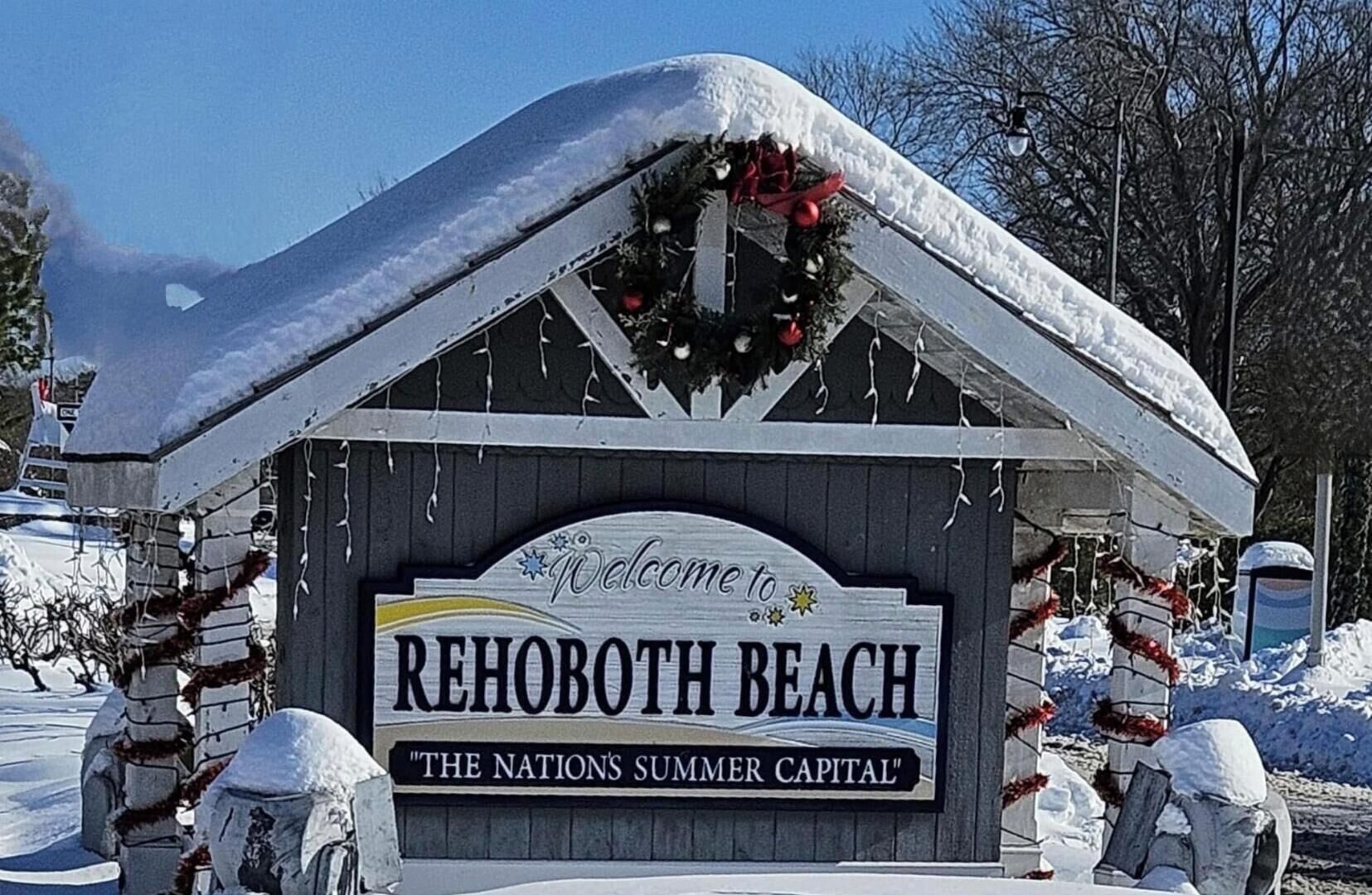 A wooden sign with "Welcome to Rehoboth Beach" and "The Nation's Summer Capital" written on it, surrounded by snow and holiday decorations.