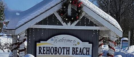 A wooden sign with "Welcome to Rehoboth Beach" and "The Nation's Summer Capital" written on it, surrounded by snow and holiday decorations.