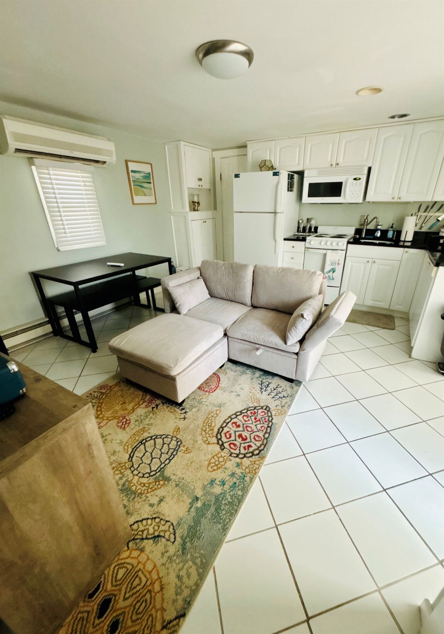 A living room with a sofa, ottoman, and a patterned rug. There is a dining table with chairs, a kitchen area with white cabinets and appliances, and a ceiling light fixture.