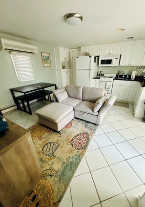 A living room with a sofa, ottoman, and a patterned rug. There is a dining table with chairs, a kitchen area with white cabinets and appliances, and a ceiling light fixture.