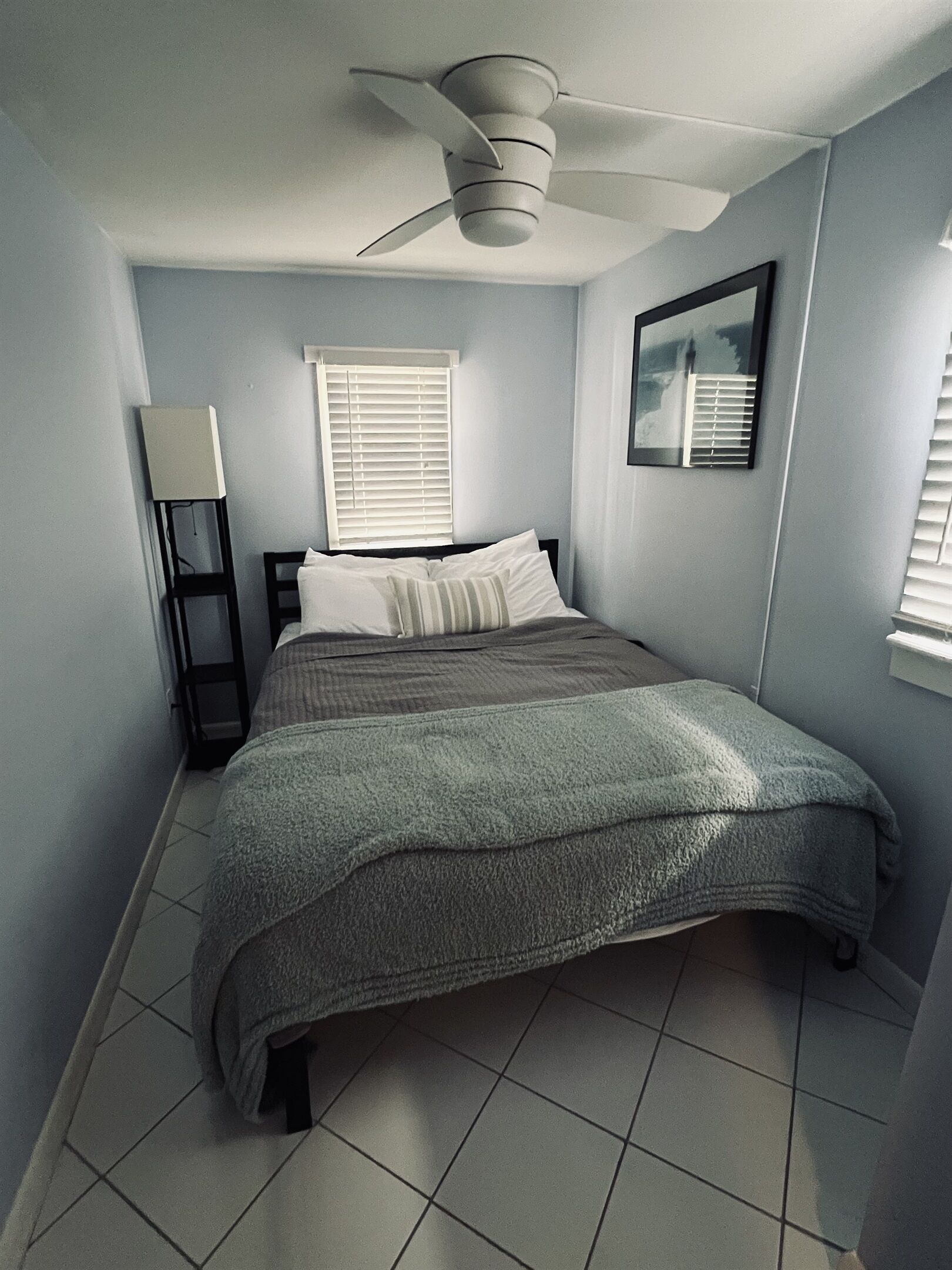 Une chambre à coucher avec un lit, un ventilateur de plafond, une fenêtre avec des stores et un tableau accroché au mur.