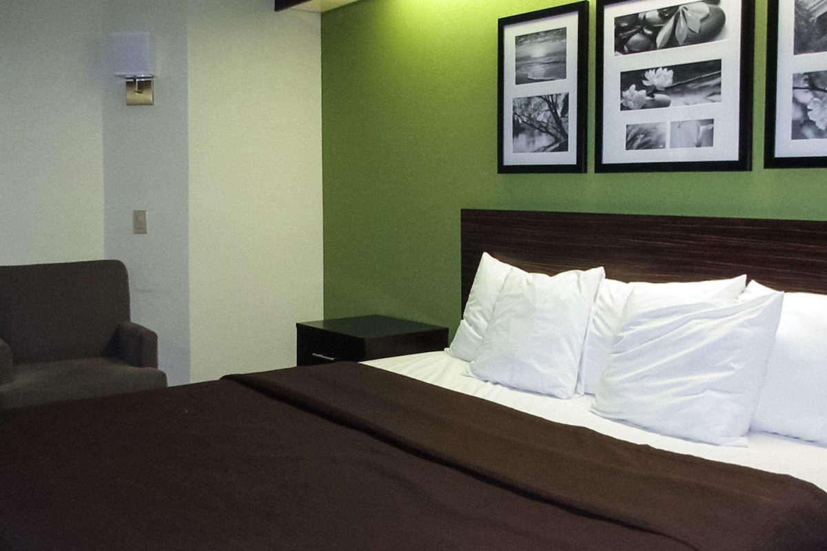 A hotel room with a bed, a nightstand, a chair, and a wall with framed pictures.