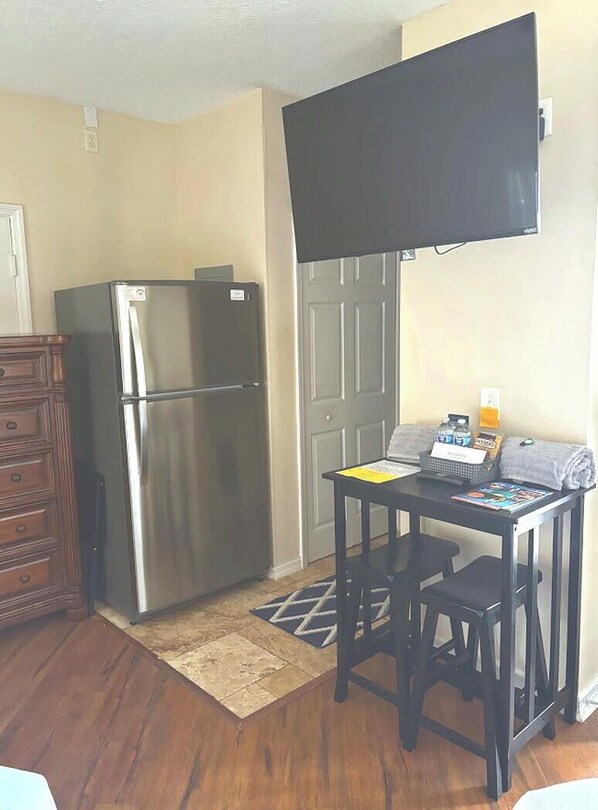 A compact kitchen with a stainless steel refrigerator, a wooden dresser, a small table with a laptop, and a flat-screen TV mounted above the table. - Right Direction Rentals Studio Villa 9897 (Port St. Lucie)