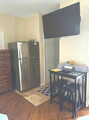 A compact kitchen with a stainless steel refrigerator, a wooden dresser, a small table with a laptop, and a flat-screen TV mounted above the table.