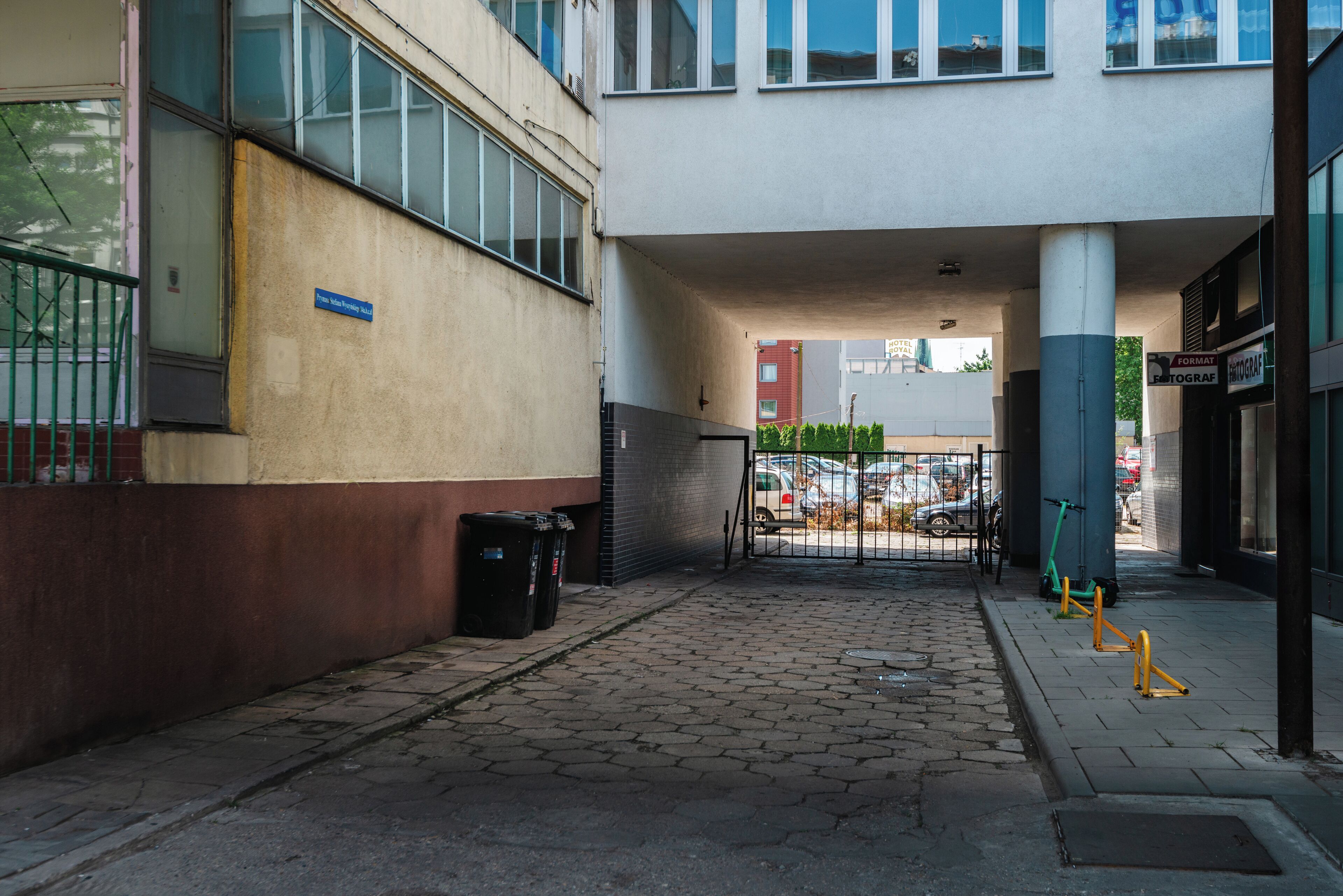 Entrada a un edificio con un camino de acceso con puerta, un contenedor de basura y una scooter estacionada afuera.