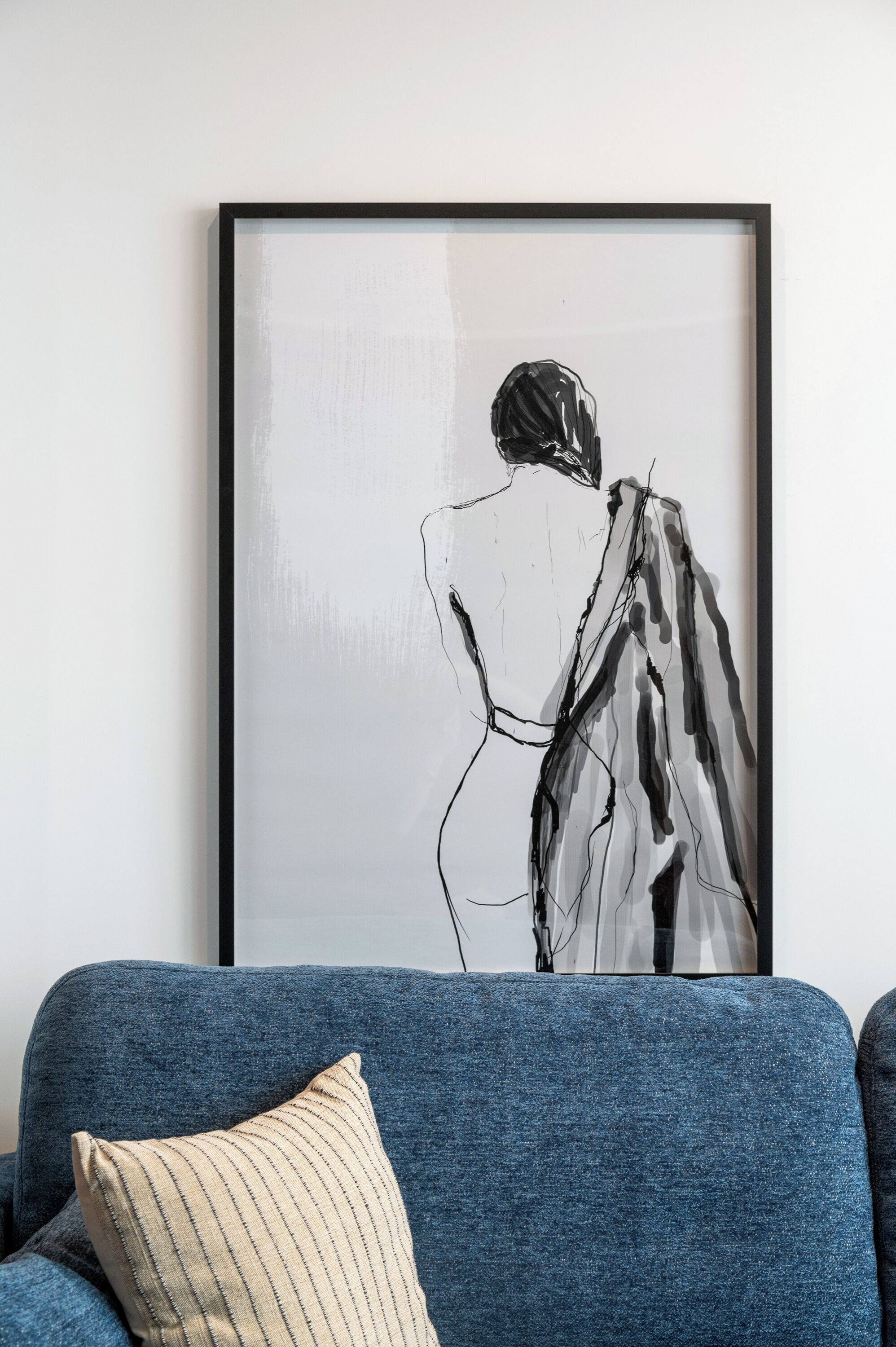 Een lijst met een zwart-wit abstract schilderij aan een witte muur, een blauwe bank met een gestreept kussen, en een donkerblauwe fauteuil.