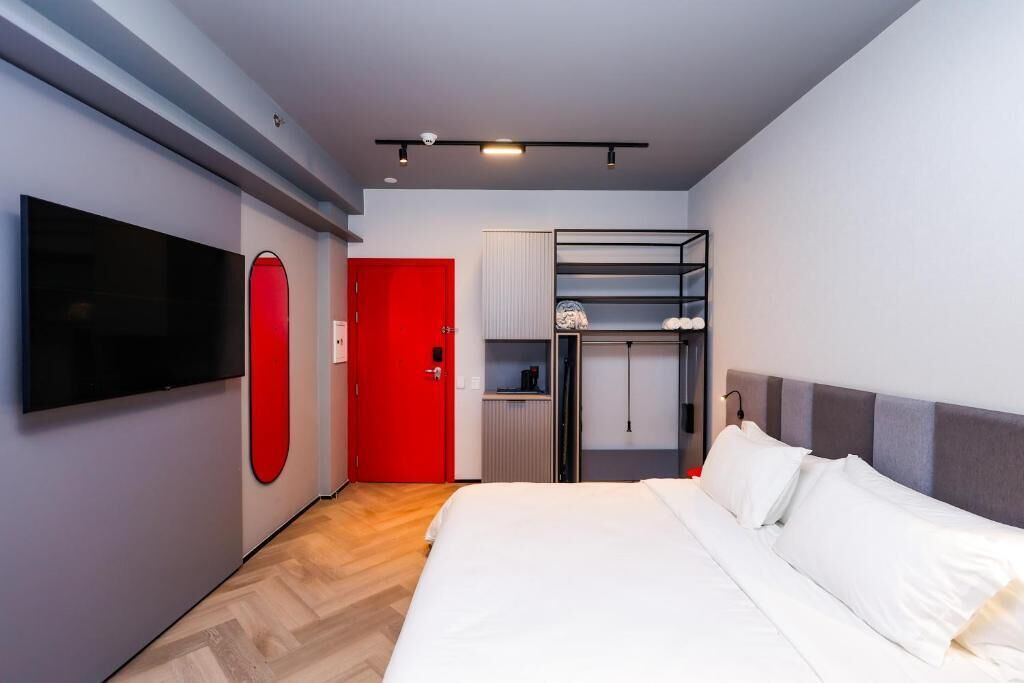 Una habitación de hotel moderna con una cama grande, un televisor de pantalla plana montado en la pared, una puerta roja y un armario empotrado con estantes.