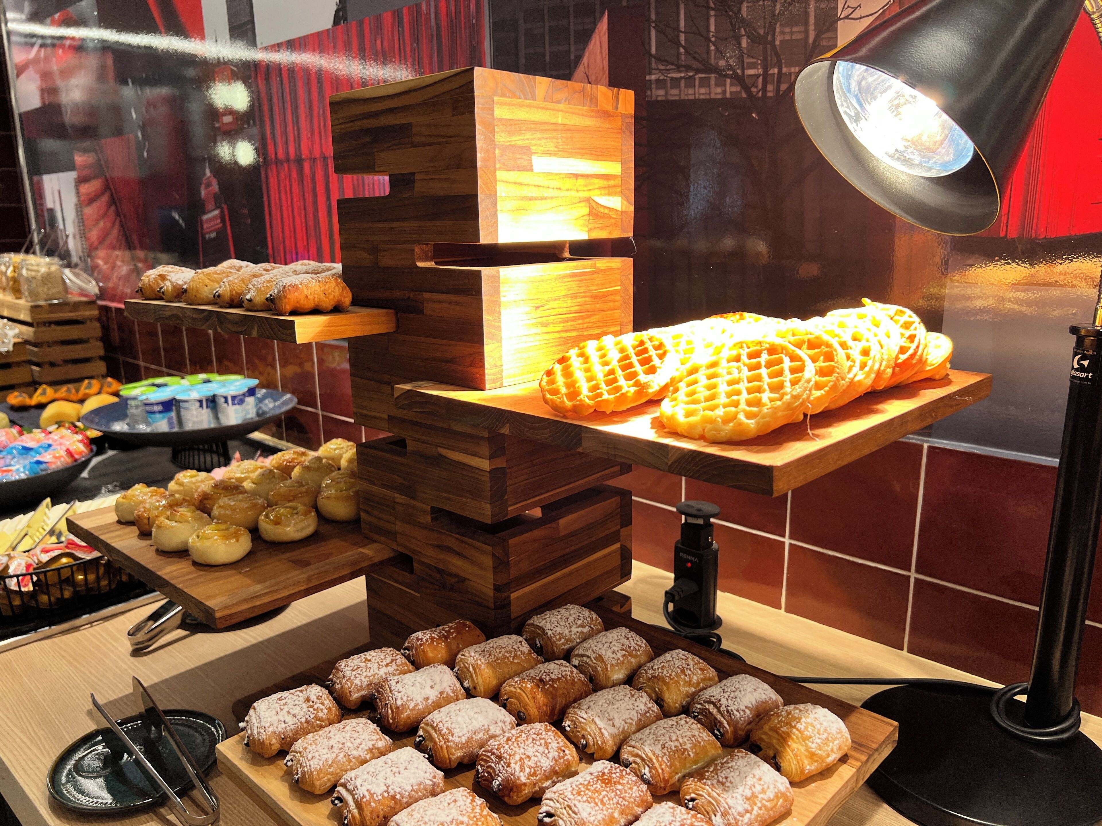 Una vitrina de pastelería con diversos pasteles y pan, incluyendo gofres y croissants, dispuestos sobre soportes de madera y una tabla de madera.