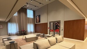 A modern living room with a sectional sofa, ottomans, and a coffee table. There is a large window with curtains, a painting on the wall, and a door leading to another room. - Radisson RED Ibirapuera (Sao Paulo)