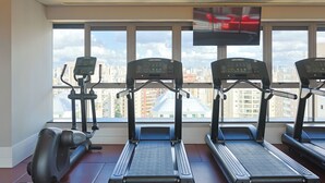 A gym with treadmills and a stationary bike, large windows with a city view, and a mounted TV. - Radisson RED Ibirapuera (Sao Paulo)