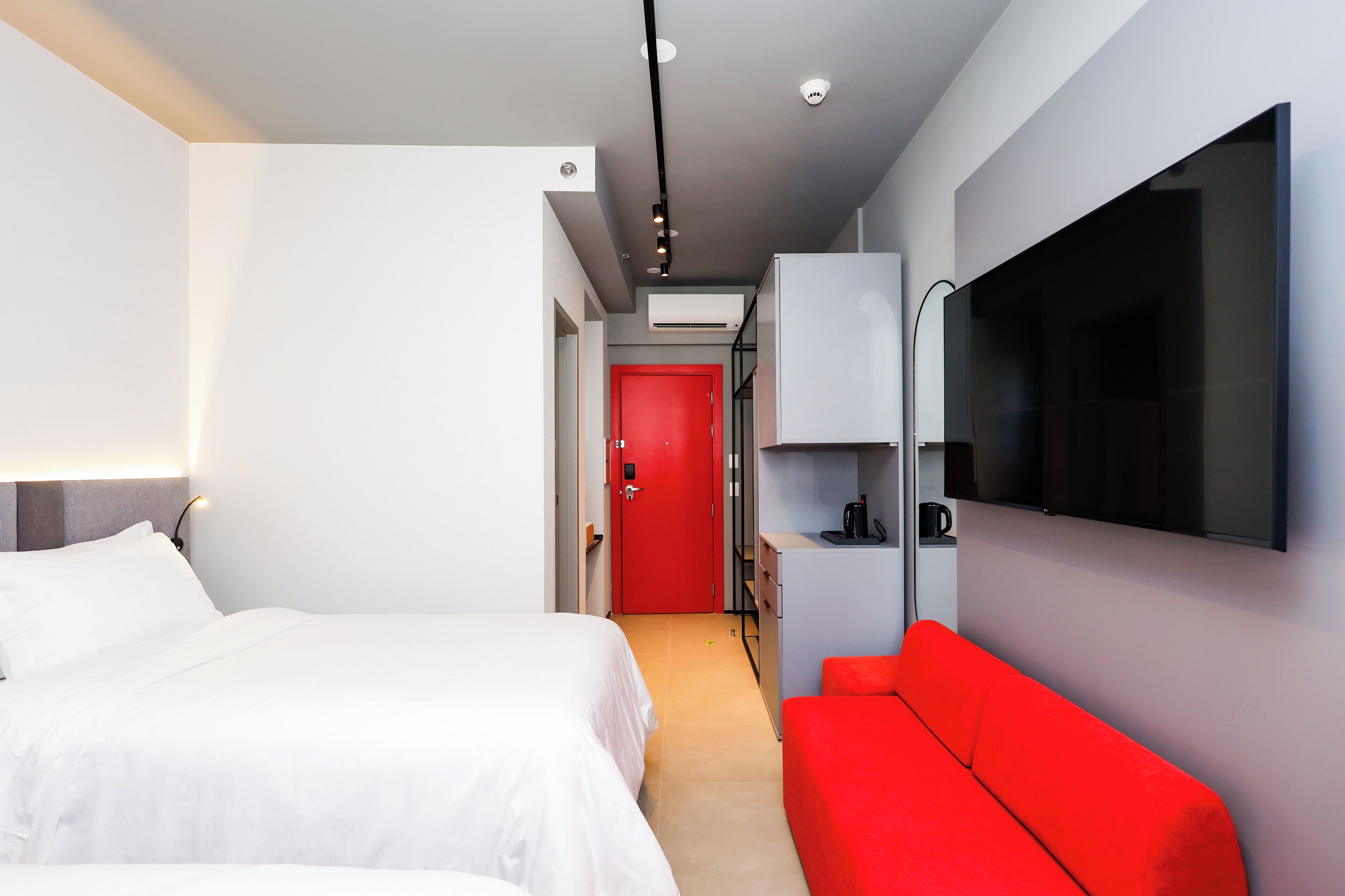 A modern hotel room with a bed, a red sofa, a TV, and a red door in the background.