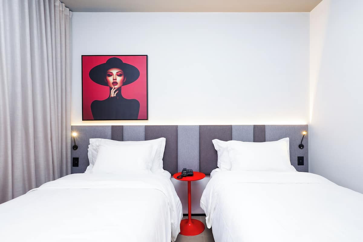 A hotel room with two beds, a red side table, a framed artwork above the beds, and a modern headboard.