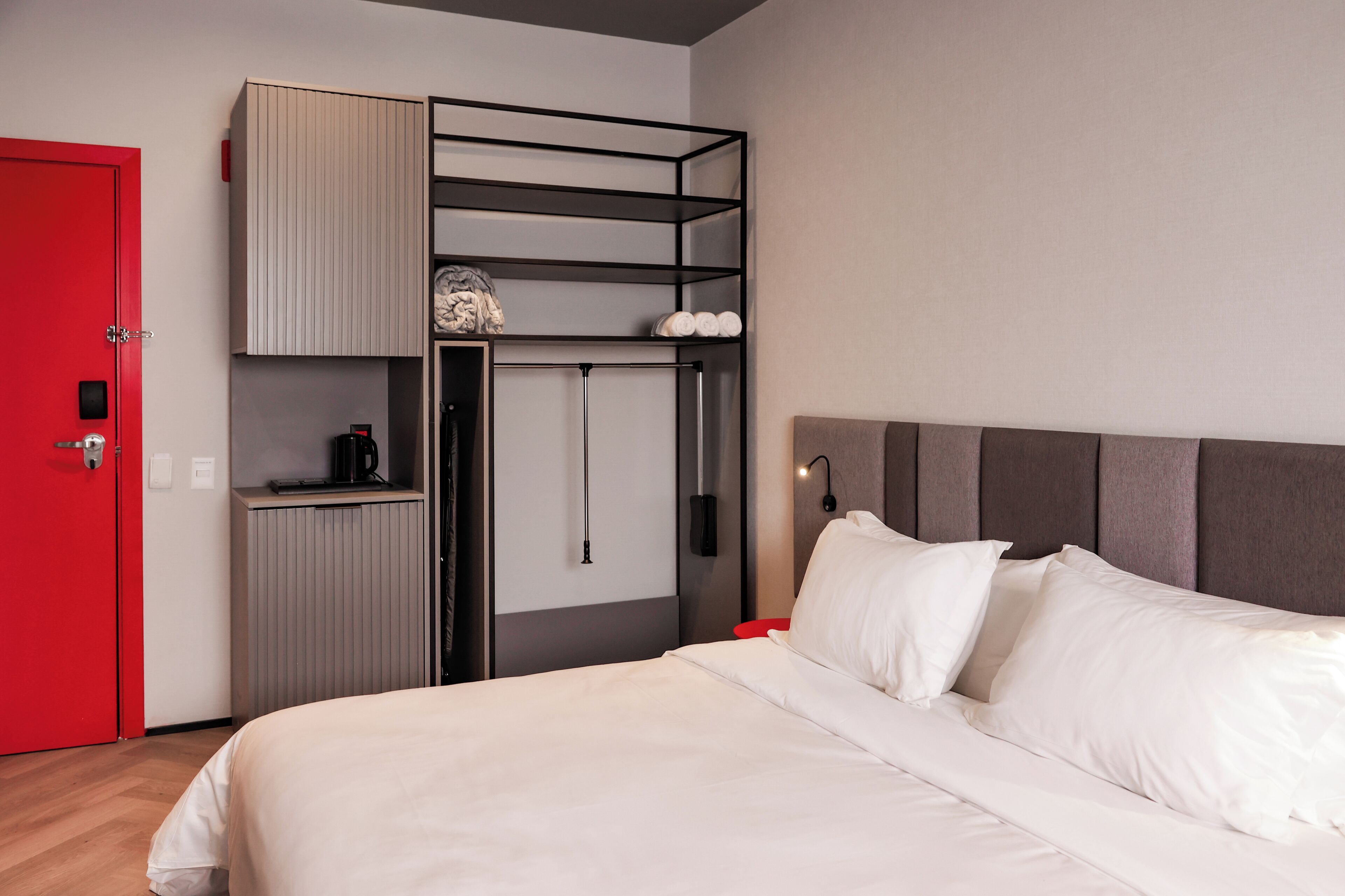 A modern hotel room with a bed, a red door, a built-in wardrobe, and a small kitchenette area.