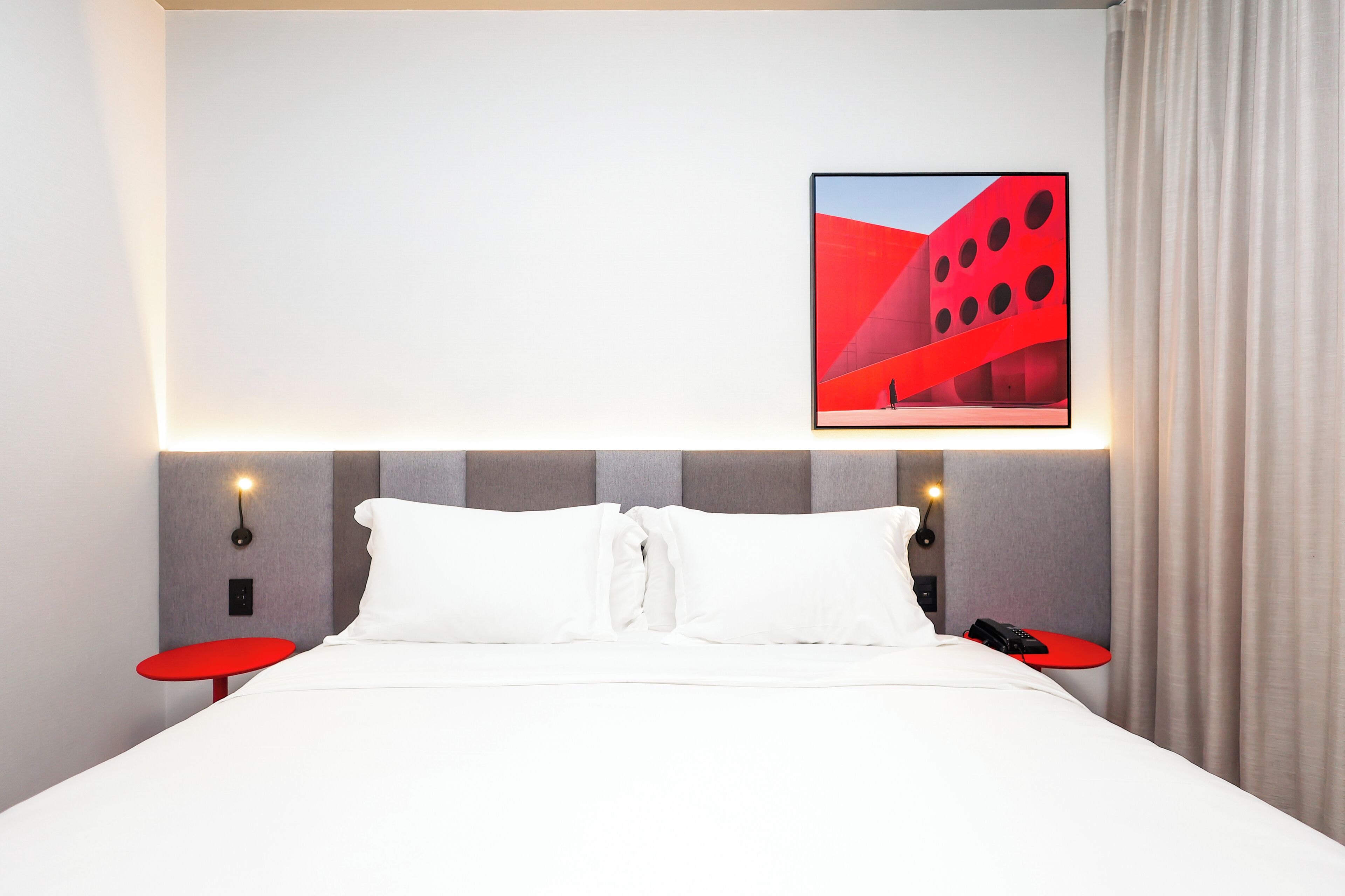 A hotel room with a bed, two red side tables, a wall-mounted light, and a framed picture above the bed.