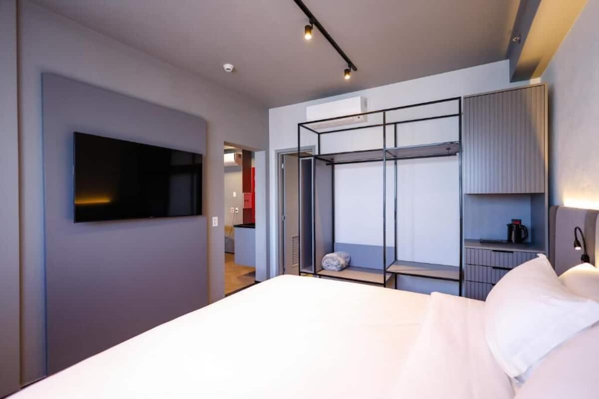 A modern hotel room with a large bed, a flat-screen TV mounted on the wall, a walk-in closet with shelves, and a small seating area with a table and chairs.