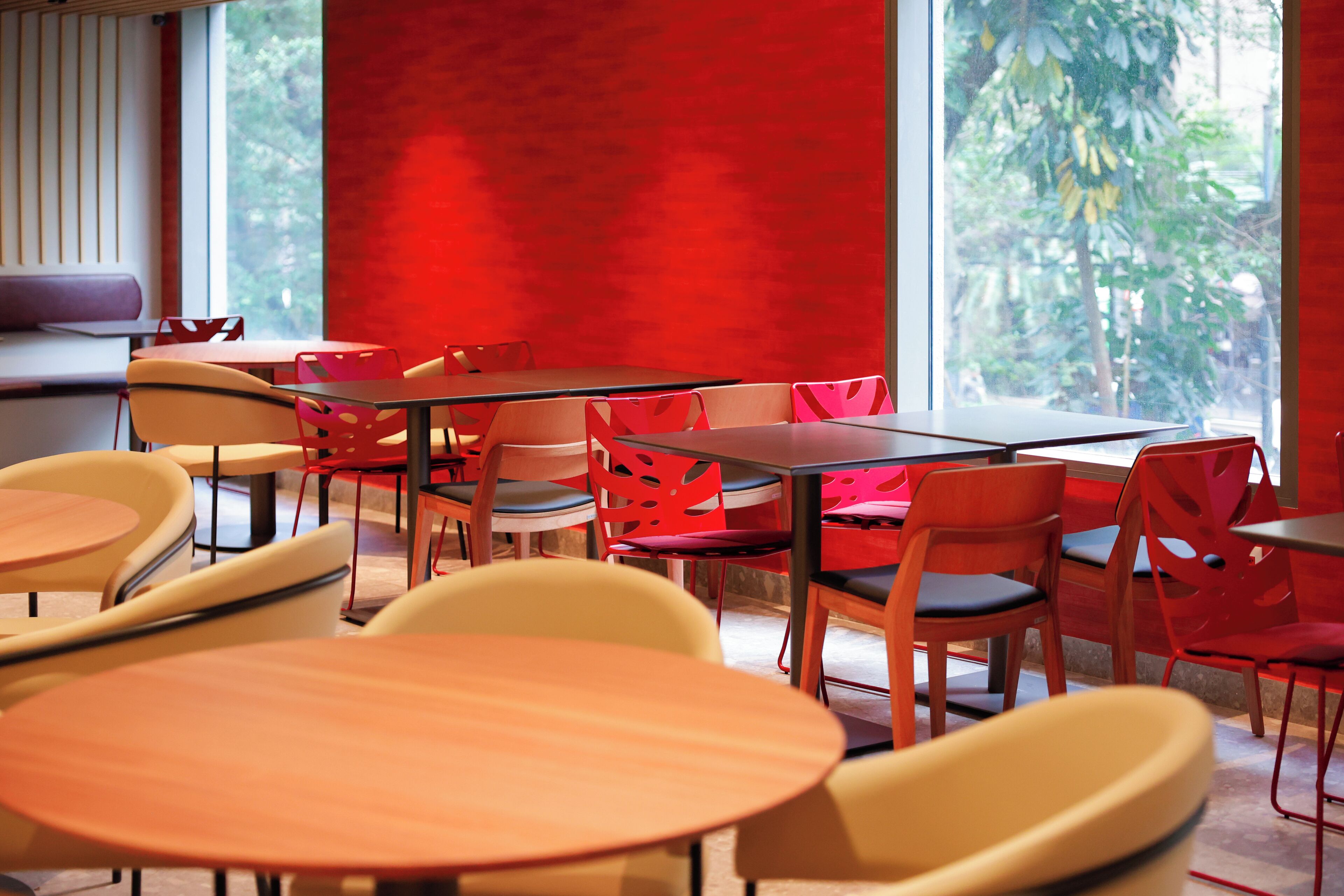 Un espace de restauration moderne avec des tables et des chaises rondes, de grandes fenêtres offrant une vue sur la verdure et un mur d’accent rouge.