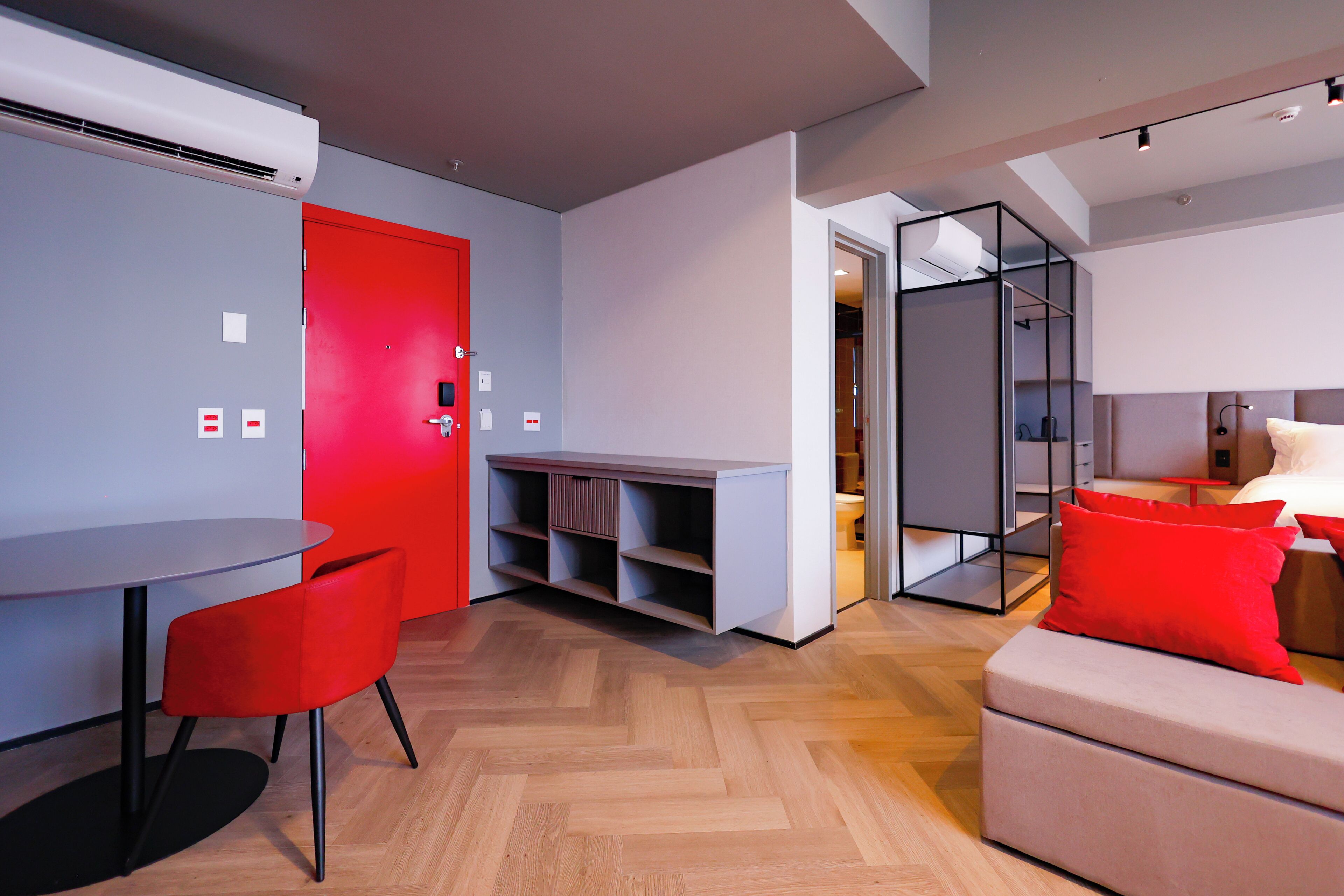 Una habitación de hotel moderna con zona de comedor, una puerta roja, un sofá con cojines rojos y un suelo de madera con patrón de espiga.