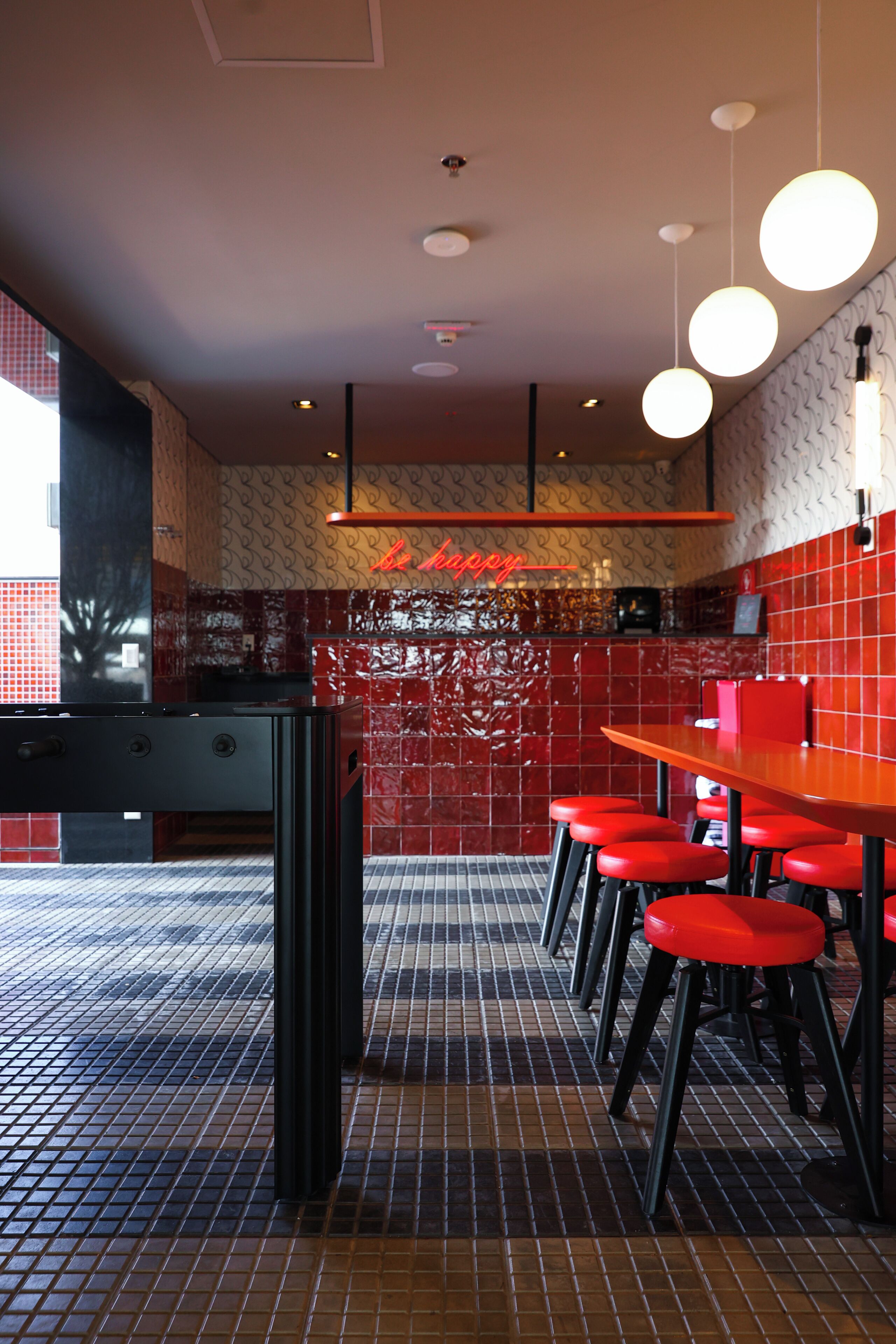 Interior de restaurante com chão de azulejos, cadeiras pretas e vermelhas, uma mesa comprida e um letreiro de neon vermelho.