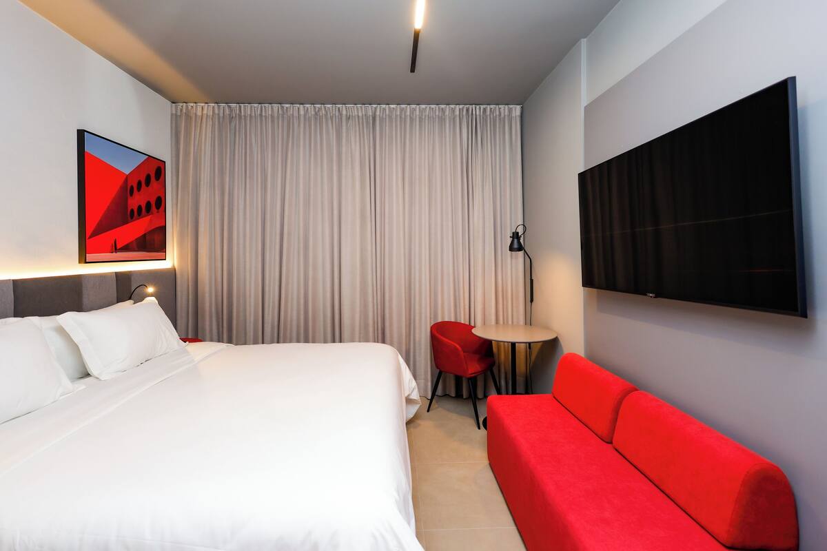 A modern hotel room with a large bed, a red sofa, a TV mounted on the wall, and a small table with a chair.