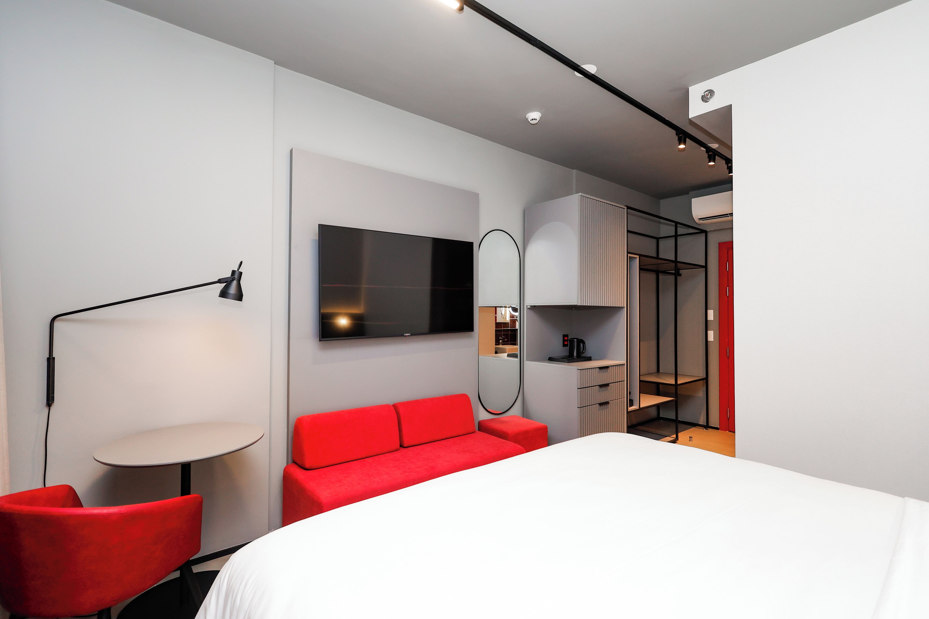 A modern hotel room with a large bed, red upholstered chairs, a round table, a TV, and a wardrobe.