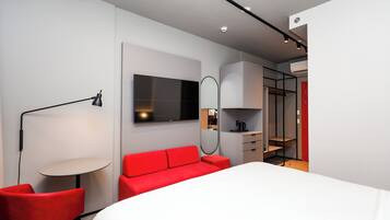 A modern hotel room with a large bed, red upholstered chairs, a round table, a TV, and a wardrobe.