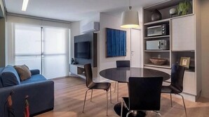 A modern living room with a sofa, a dining table with chairs, a TV, and built-in shelves with kitchen appliances. - Urban Home Stay - Santa Maria (Recife)