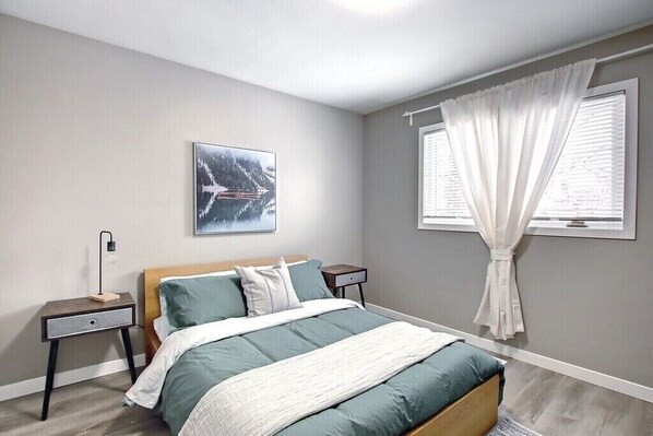 A bedroom with a bed, nightstands, a wall-mounted lamp, a window with white curtains, and a framed picture on the wall. - Historical Highlands Home (edmonton)
