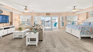A spacious living room with a large window, a ceiling fan, and a bed with a floral bedspread. - Seaside Escape Mansion (Ocean City)