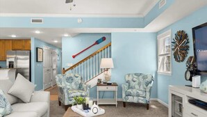 A living room with a light blue wall, a wooden staircase, and a kitchen area with wooden cabinets and a stainless steel refrigerator. - Seaside Escape Mansion (Ocean City)