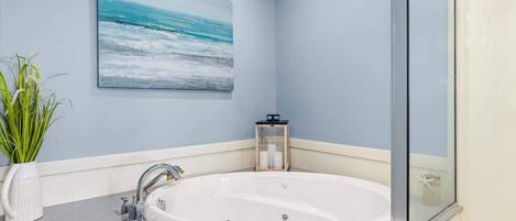A bathroom with a large bathtub, a sink, a glass shower enclosure, a potted plant, and a painting of the ocean on the wall.