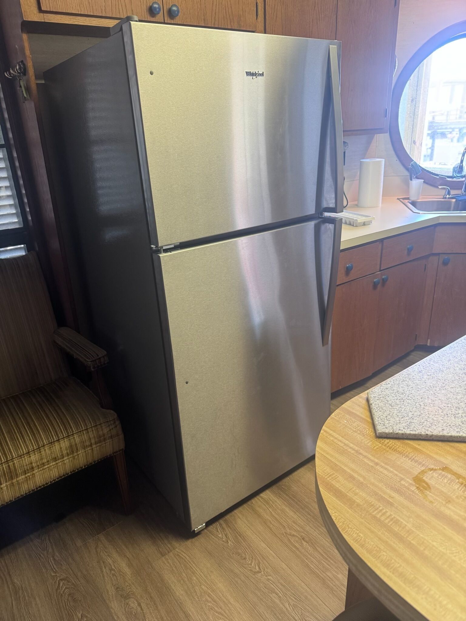 Eine kompakte Küche mit Edelstahlkühlschrank, Holzschränken, einem kleinen Esstisch und einem Fenster mit Jalousien.