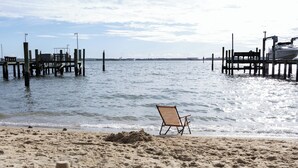 A beach with a wooden chair and a sandcastle, a pier with boats, and a clear sky.
