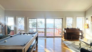 A spacious room with a pool table, a piano, and a view of the water through large windows.