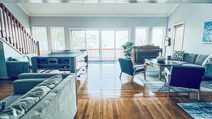 A spacious living room with a wooden floor, a pool table, a sofa, a dining table, and a piano. There are large windows and a staircase.