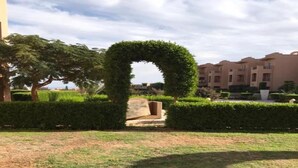 A landscaped area with a circular hedge archway, a stone bench, and a building in the background.
