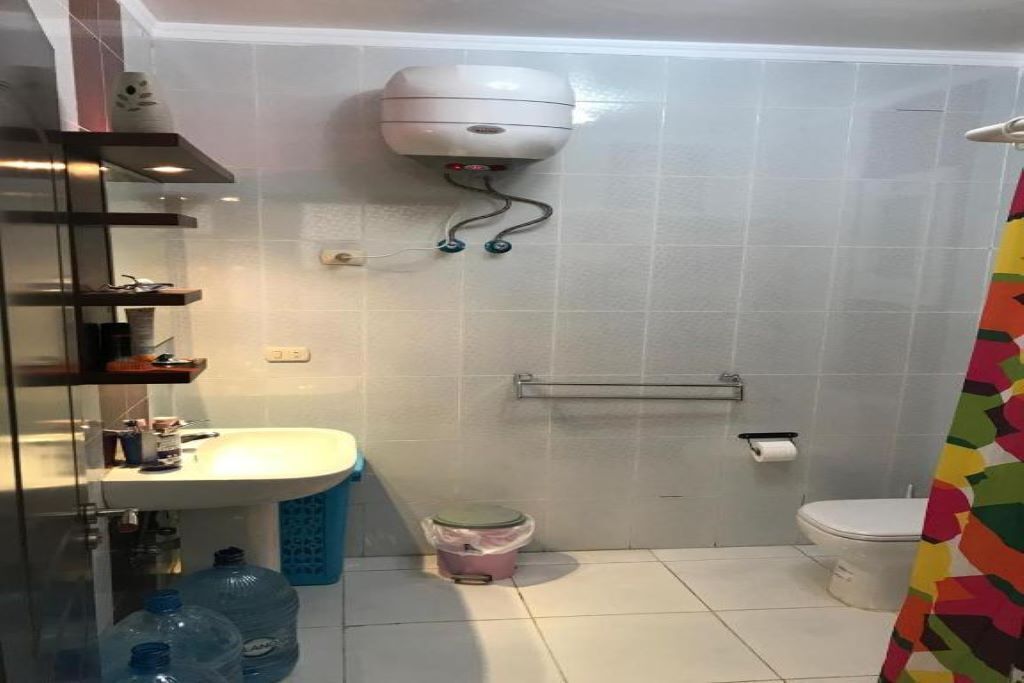 A bathroom with a white sink, toilet, and a wall-mounted toilet paper holder. There is a trash bin and a water dispenser on the wall.