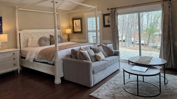 A four-poster bed with a grey upholstered sofa and a round coffee table in a well-lit room with a large window. - Luxury Lodging, Lakefront View, King Bed w/Private Bath, Private Patio. (Elizabethtown)