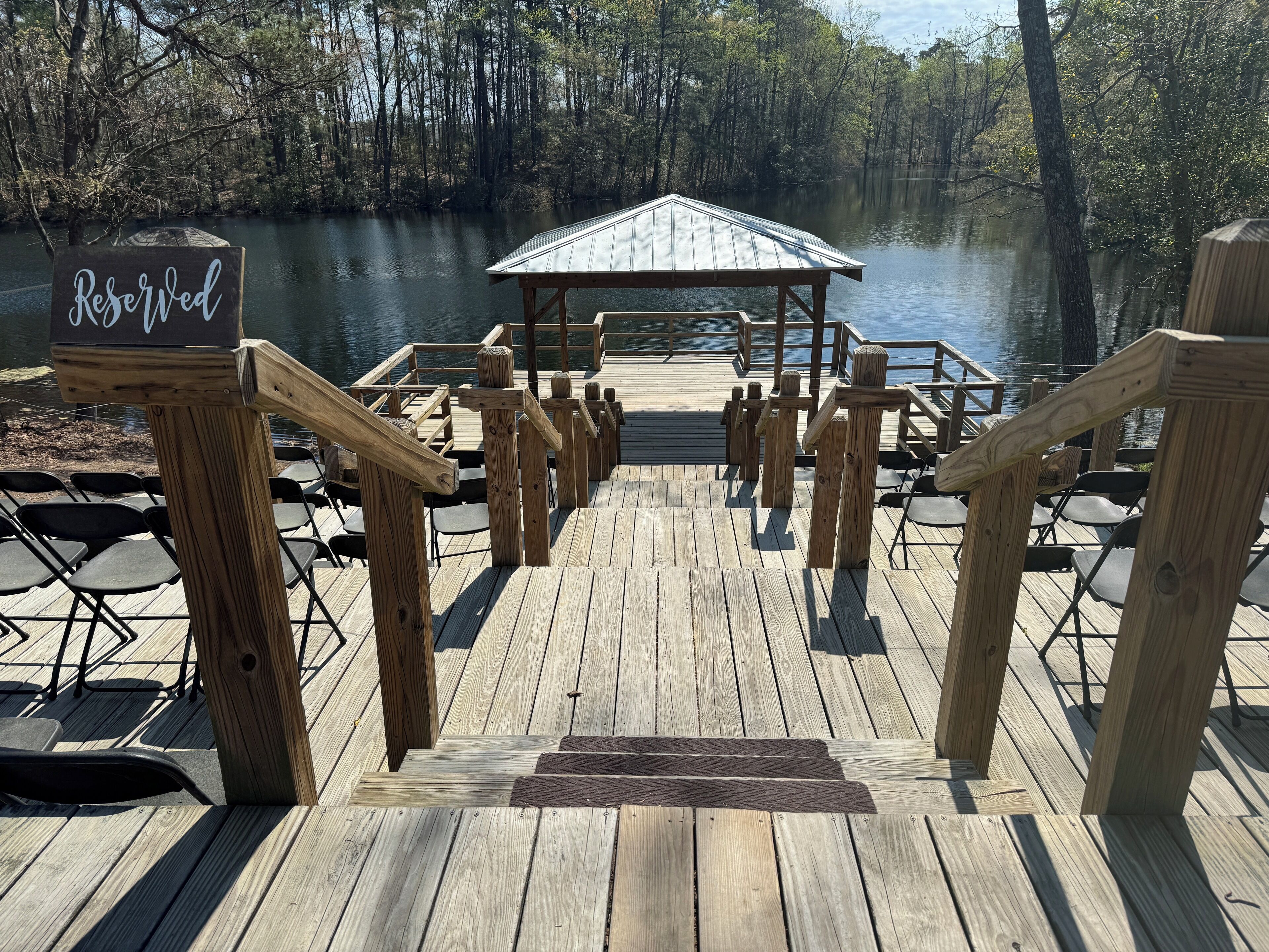 A wooden dock leading to a gazebo by a calm lake, with chairs and a 'Reserved' sign.