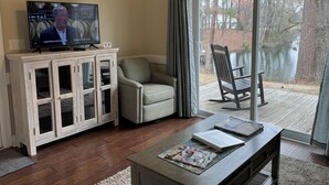 Un salon comprenant un téléviseur, un meuble en bois, un fauteuil rembourré, une table en bois avec des magazines, et une vue sur un lac à travers de grandes portes vitrées.