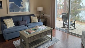 Una sala de estar con un sofá azul, una mesa de centro con revistas, una lámpara y vista a un lago a través de una puerta de cristal.