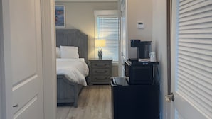 Um corredor estreito com uma cama, mesa de cabeceira, abajur e um pequeno armário preto.