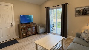 Un salon doté d’un ventilateur de plafond, d’un téléviseur posé sur un meuble en bois, d’un canapé et d’une porte-fenêtre coulissante donnant sur un espace extérieur.