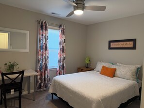 A bedroom with a bed, a nightstand, a chair, a window with curtains, a ceiling fan, and a wall with a framed quote.