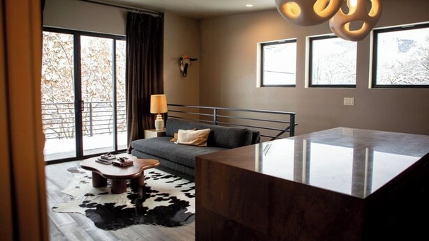 Ein modernes Wohnzimmer mit einer Couch, einem Couchtisch und einem Glastisch. Große Fenster mit Vorhängen, eine Lampe und ein an der Wand befestigter Tierkopf sind zu sehen.