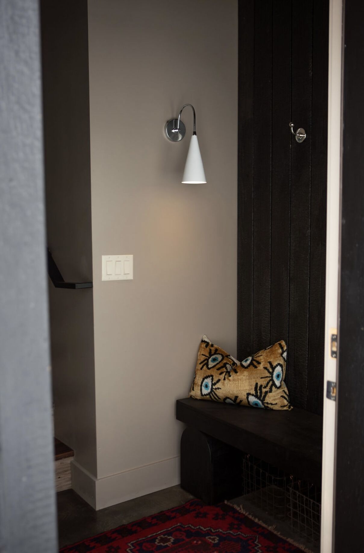 A hallway with a bench, a patterned pillow, a wall-mounted light fixture, and a red patterned rug.