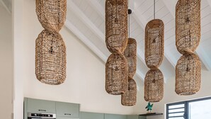 A modern kitchen with a central island, hanging woven light fixtures, and a view through a window.