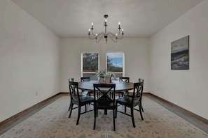 A dining room with a round table, six chairs, a chandelier, two windows with blinds, and a painting on the wall. - Luxury in the Hill Country, 10-mins to Downtown! (Fredericksburg)