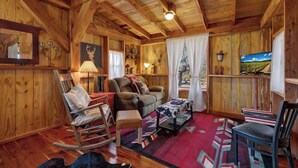 Ein rustikales Holzinterieur mit Schaukelstuhl, Sofa, Hocker, Fernseher und einem Fenster mit Blick auf einen Weinberg.