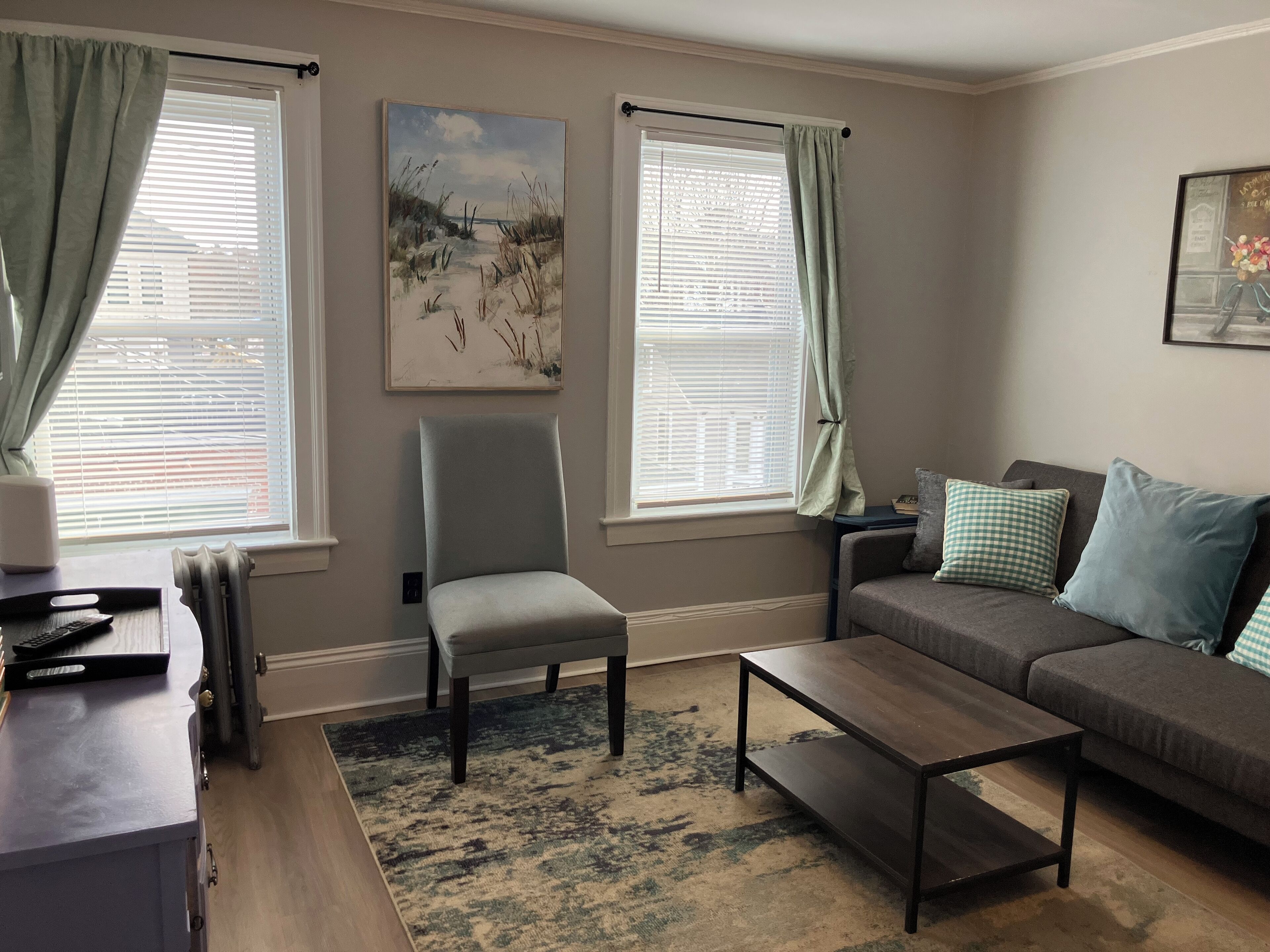 Una sala de estar con un sillón gris, una mesa de centro de madera oscura, un sofá gris con cojines azules y un cuadro en la pared.